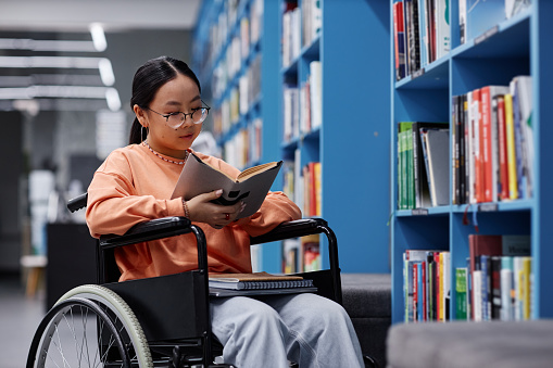 Portrait of Asian young woman with disability reading book in library, student inclusivity concept