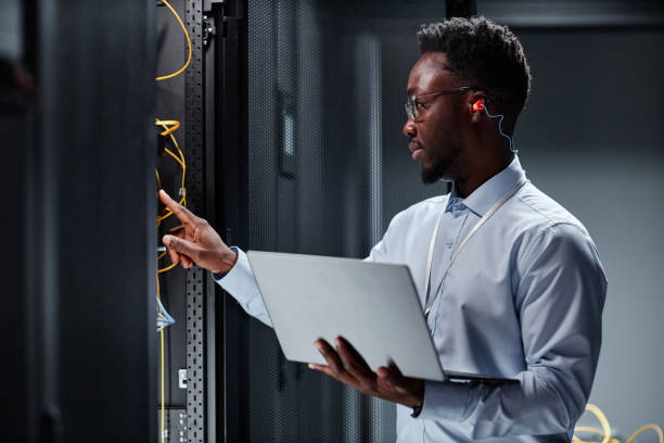 jeune ingénieur réseau travaillant avec des serveurs dans un centre de données et tenant un ordinateur portable - network server data center installing photos et images de collection