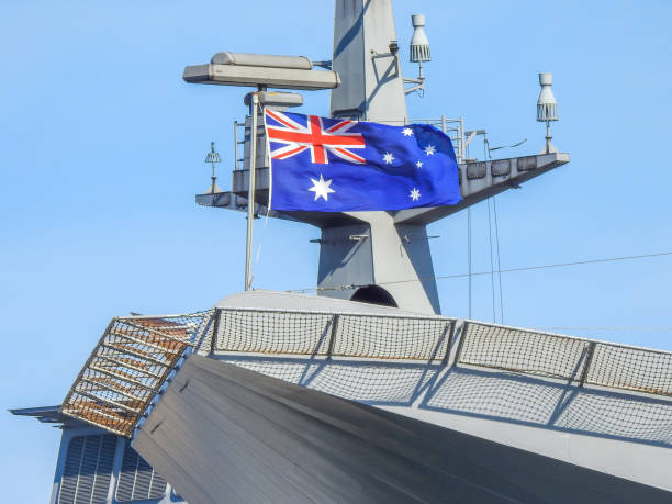HMAS Adelaide National Flag An Australian flag flies on the starboard corner of flight deck of HMAS Adelaide, docked at Garden Island, Sydney Harbour.  She is one of two Canberra-class Amphibious Assault Ships of the Royal Australian Navy.  In the background is the mast of HMAS Supply.   This image was taken from Mrs Macquarie's Chair on a windy and sunny afternoon on 14 January 2023. australian navy stock pictures, royalty-free photos & images