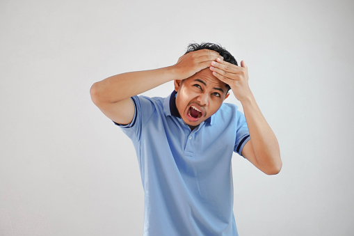 Asian man head in pain, holding head with both hands wearing blue t shirt isolated on white background