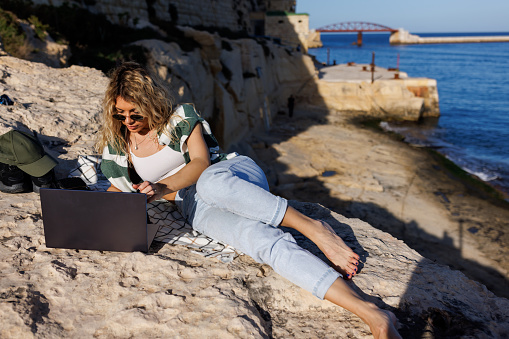Young woman lying on side and using laptop on rocky beach on sunny day