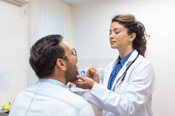 un jeune homme est assis sur une table d’examen en face de son médecin. le médecin tend la main vers l’avant avec un abaisse-langue alors que l’homme lève les yeux et tire la langue. - doctor patient male tongue depressor photos et images de collection