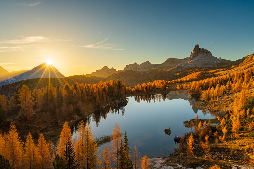 Star shape sunrise over mountain peak at calm lake high up in the dolomites with orange colored larch trees in autumn