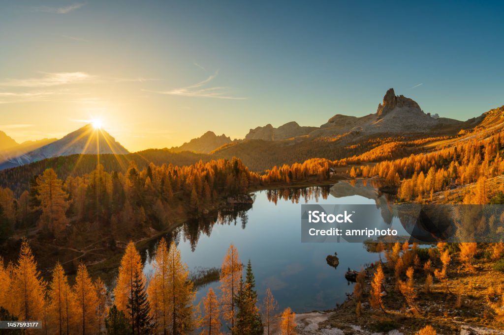 Breathtaking morning at mountain lake with sun rising Star shape sunrise over mountain peak at calm lake high up in the dolomites with orange colored larch trees in autumn Awe Stock Photo