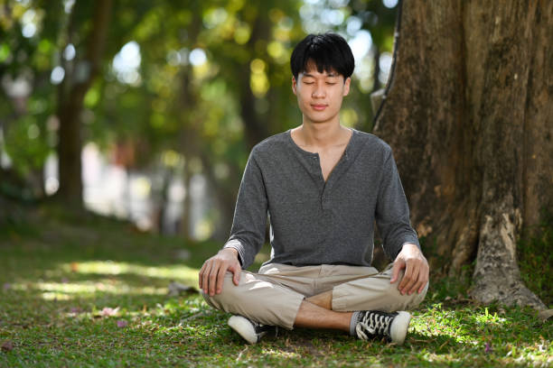 Pleasant asian man sitting in lotus pose sitting on green grass. Meditation and mental health concept. stock photo