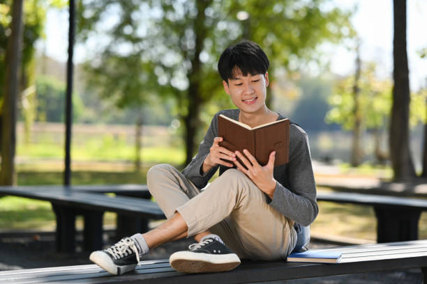 istock Young asian man student reading interesting book while sitting on bench in city park. Education, lifestyle concept. 1457595968