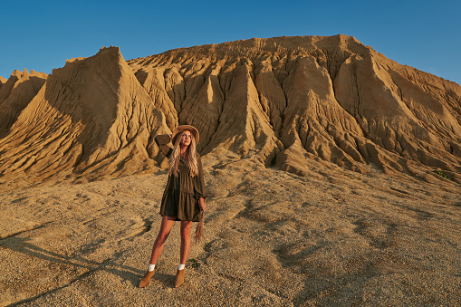 a beautiful young woman in a dress and a hat with dry grass in her hands on the background of a huge sandy mountain and a beautiful clear blue sky. High quality photo