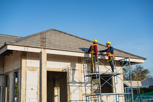 Two men wearing protective clothing installing solar panels on the roof of a newly built house at a village construction site.