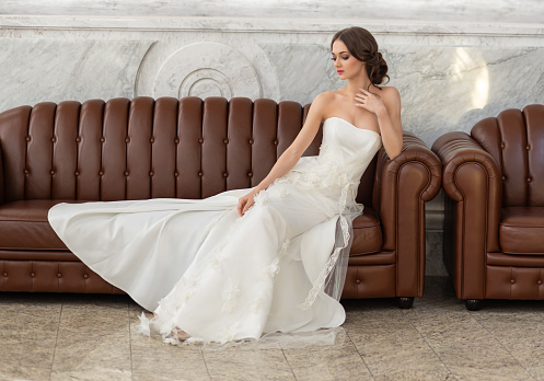 Молодая хрупкая брюнетка. Невеста в роскошном свадебном платье сидит на диване в красивом солнечном свете из окна. Свадебная прическа и макияж. Фото для  салона красоты
