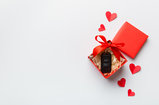 Black car key in a present box with a ribbon and red heart on colored background. Valentine day composition Top view.