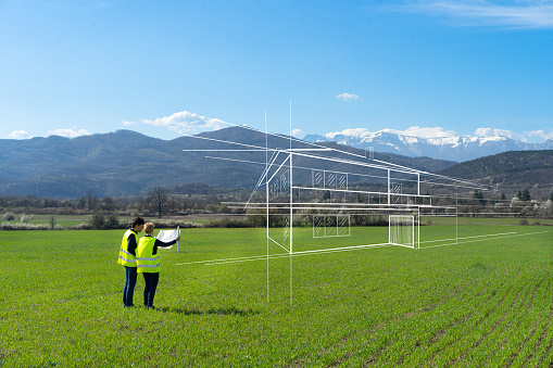 A couple are standing in a large field with hills and mountains in front of them as they hold up their house plans and try and imagine their new property, with a drawing of a house imposed onto the picture