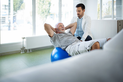 Closeup side view of a mature man having medical condition evaluation at doctor's office. He's performing crunches on a Pilates ball.
