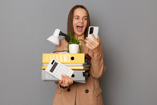 Portrait of extremely happy young pretty woman wearing beige jacket holding documents and her office stuff posing isolated over gray background, holding smart phone, expressing positiveness.