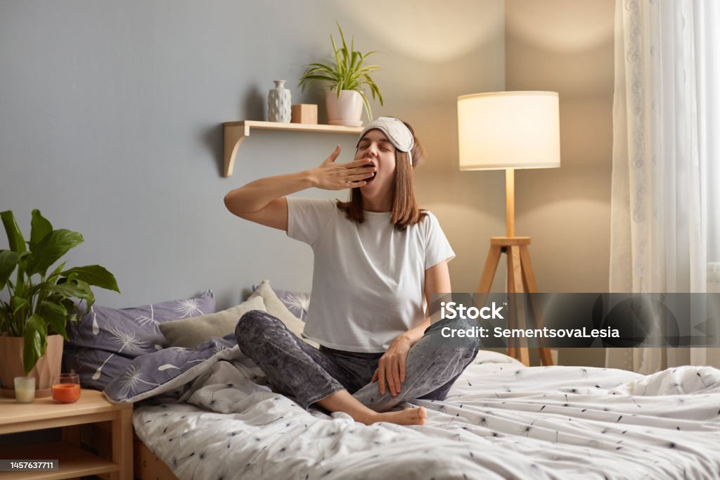 Portrait of sleepy tired woman wearing sleeping mask sitting on bed in cozy bedroom, yawning and covering mouth with palm, feels sleepy, wants to have nap. Yawning Stock Photo