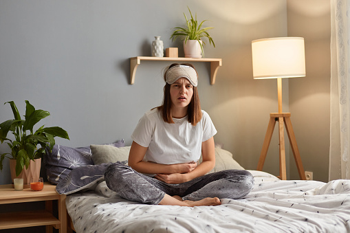 Horizontal shot of sick unhealthy sad woman wearing sleeping mask sitting on bed in cozy bedroom, having terrible strong stomachache, frowning face from pain.