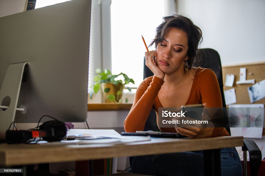 Bored young woman sitting at her desk, anticipating a text message Portrait of annoyed young woman sitting with head in hand at her desk in a small office, holding her smart phone and waiting to receive a text message, distracted from work. Frustration Stock Photo