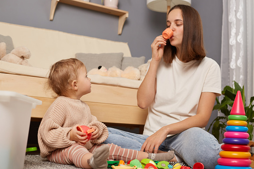 Portrait of dark haired mother and daughter playing with toys on the floor, mother holding tiny colorful cup, pretended to drink, kid looking at mommy with curious expression.