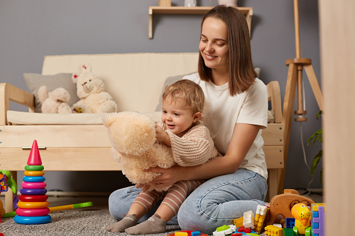 Indoor shot of dark haired woman wearing white T-shirt sitting on floor near sofa and holding her baby daughter, playing with soft toy teddy bear, expressing positiveness.