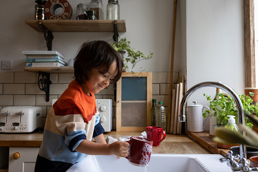 A three-quarter length shot of a male child washing a red mug at a sink at home. He is smiling and concentrating on what he is doing wearing casual clothing.