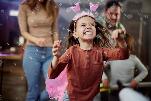 Joyful little girl having fun while catching bubbles on a party at home. There are people in the background.