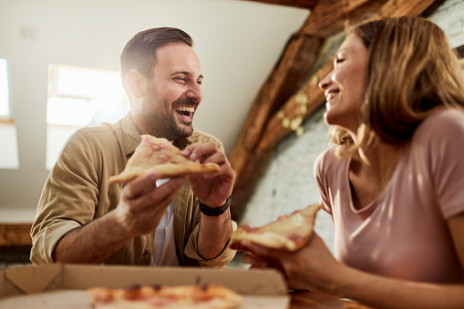 Young happy couple communicating while having pizza for lunch in dining room. Focus is on man.