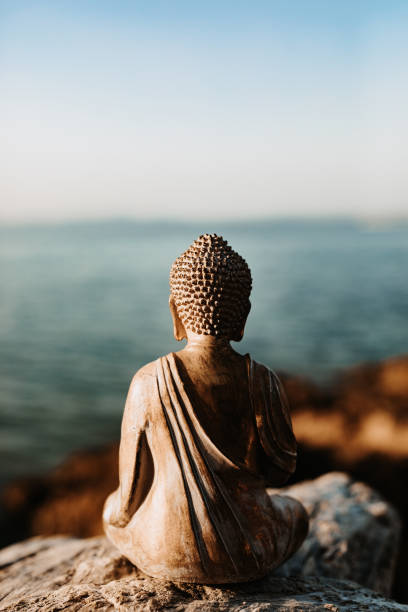 alte buddha-figur von hinten, die auf einem stein vor dem meer sitzt - buddha image stock-fotos und bilder