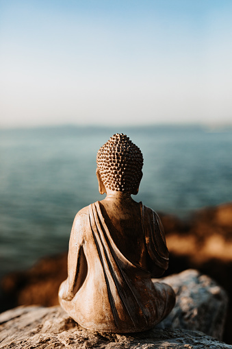 Old Buddha figure from behind sitting on a stone in front of the sea. Selective focus. Part of a mindfulness series.