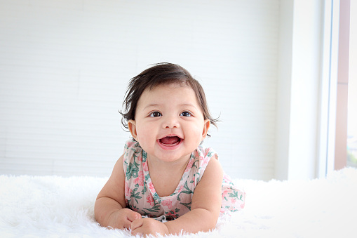 Portrait of six months crawling baby on fluffy white rug, happy smiling adorable sweet little girl kid lying on bed in bedroom, childhood and baby care concept