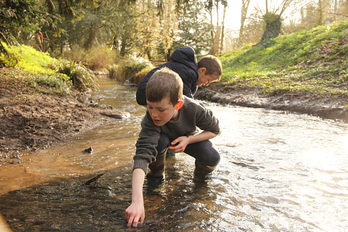 Two young boys exploring in a stream during winter, they are wearing warm clothing and putting their hands in water