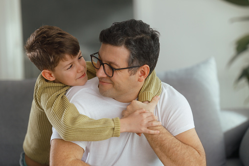 Shot of a cute teenage boy hugging his father on the sofa at home. They are smiling and looking happy.