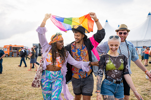 Group of friends having fun at a festival in Northumberland, North East England. They are walking side by side holding a rainbow flag above their head.