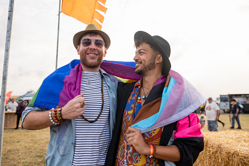 LGBTQI+ couple having fun at a festival in Northumberland, North East England. They are walking side by side with their arms around each other and a rainbow flag around their shoulders. One of the men is looking at the camera.