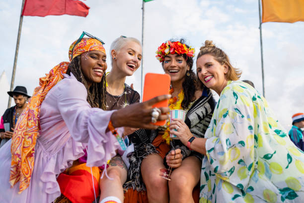 Posting to Social Media Group of female mixed age and ethnic friends having fun at a festival in Northumberland, North East England. They are laughing and drinking together, taking selfies on a mobile phone. festival goer stock pictures, royalty-free photos & images
