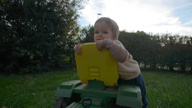 Sweet toddler runs on green lawn to sit on tractor toy