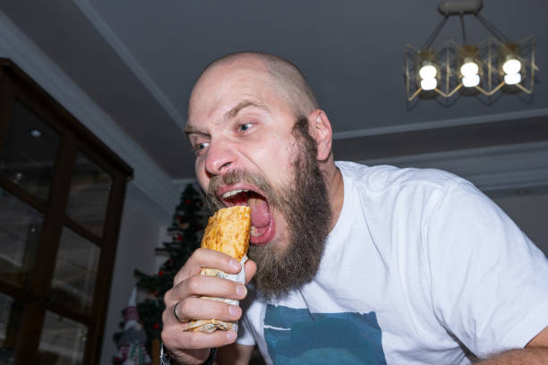 uomo dipendente che mangia fast food a casa disordinata. ragazzo molto affamato. concetto di dieta. concetto divertente di cibo malsano, snack, cibo di strada e dipendenza. - white food and drink industry hamburger cheeseburger foto e immagini stock