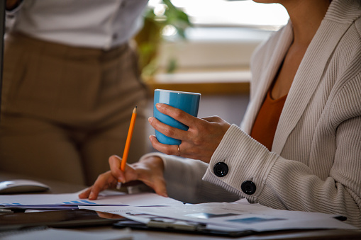 Close up shot of unrecognizable young businesswoman sitting at her desk, showing data, on computer screen, to her female colleague while having coffee together.