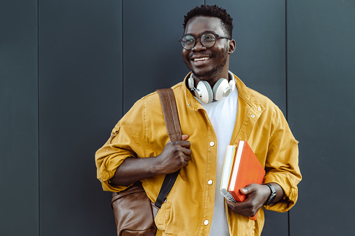 Photo of a cheerful African American young man in a yellow jacket with headphones and backpack. He is holding his notebooks.