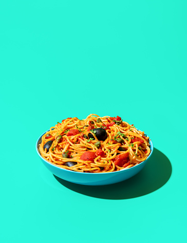 Single bowl with pasta puttanesca minimalist on a green table. Vegan italian dish, spaghetti with tomato sauce, black olives and capers.