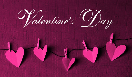 Pink paper cut hearts hanging on a clothesline with wooden clothespins. VALENTINE'S DAY text is in the center. Can be used as a design for Valentine's day holiday greeting cards or posters.