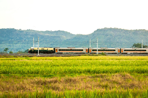 Central Java, Indonesia - August 22, 2022: An Indonesian railway locomotive GE U18C with a 1980s livery pulls Sancaka train rides on a curved railroad that passes through ricefields and mountains.