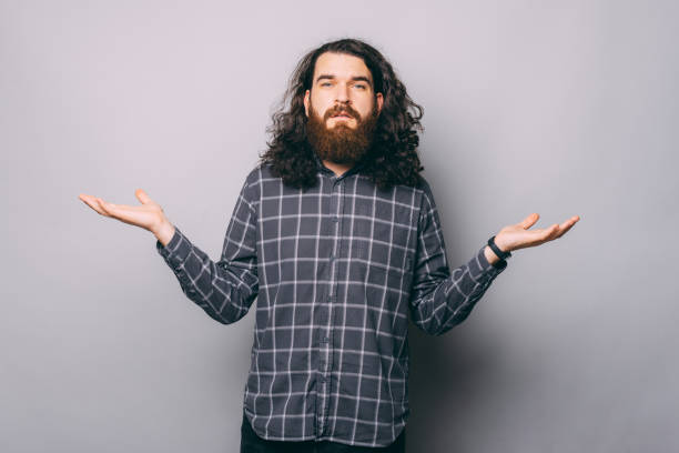 Thoughtful young bearded man standing and holding copyspace on both palms over gray background stock photo