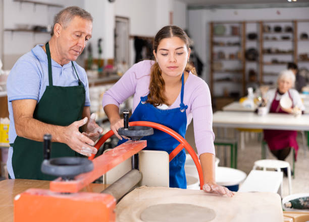 istock Elderly potter teaches woman how to roll clay on a craft machine clay press roller 1457673897