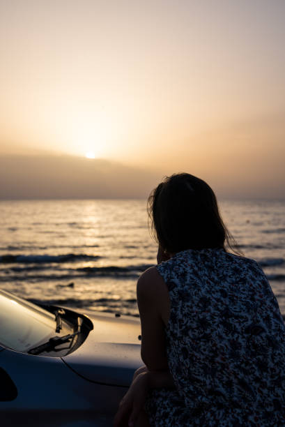 istock Woman on beach by a car looking at view with sunset vertical image 1457674007