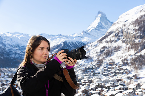Traveler photographer woman with camera taking pictures of landscape of Swiss Alps on sunny winter day