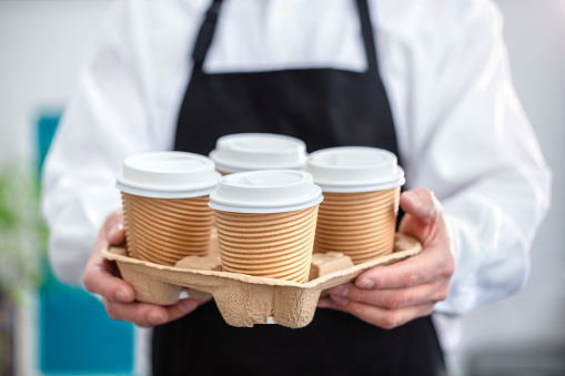 Waiter barista holding and serving take out a paper disposable cup of hot coffee in cafe