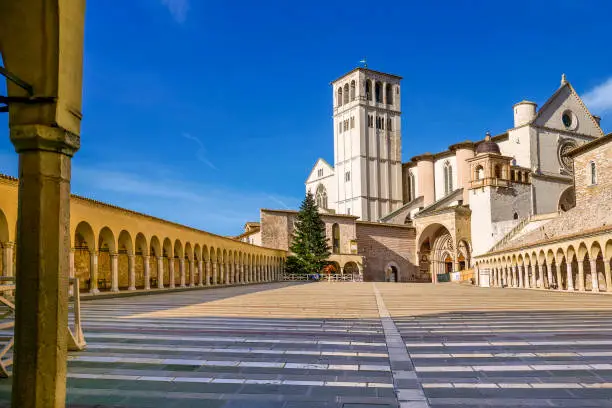 A wide angle scene of the Lower Square of the Basilica di San Francesco (Basilica of St Francis) in the medieval town of Assisi, in Umbria. On the center, the bell tower and the facade in Italian Gothic style of the Basilica dedicated to the Saint of the Poor. Built in the Italian Gothic style starting from 1228 and completed in 1253, the Basilica, which preserves the mortal remains of the Saint of the Poor from 1230, is composed of the Basilica Inferiore (Lower Basilica) and the Basilica Superiore (Upper Basilica), perfectly integrated. Over the centuries Assisi and the spirituality of its sacred places have become a symbol of peace, a point of reference for tolerance and solidarity between peoples and between the different confessions of the world. The Umbria region, considered the green lung of Italy for its wooded mountains, is characterized by a perfect integration between nature and the presence of man, in a context of environmental sustainability and healthy life. In addition to its immense artistic and historical heritage, Umbria is famous for its food and wine production and for the high quality of the olive oil produced in these lands. Since 2000 the Basilica and other Franciscan sites of Assisi have been declared a World Heritage Site by UNESCO. Image in high definition format.