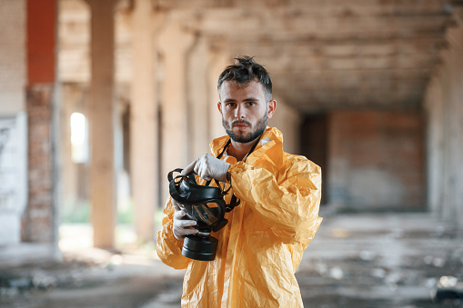 Holding gas mask. Man dressed in chemical protection suit in the ruins of the post apocalyptic building.