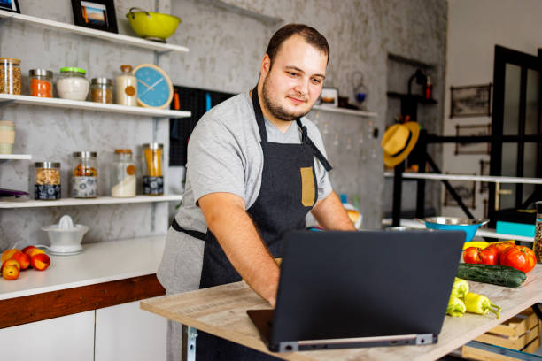 istock Overweight man using laptop while chopping vegetables in the kitchen 1457690978