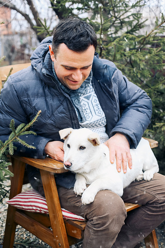 Smiling young man sitting on wooden chair with dog on laps, drinking hot aromatic grog, relaxed happy guy in warm clothes resting in backyard of private house during winter holidays. Selective focus