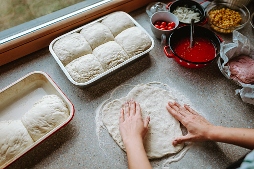 Woman preparing pizza dough at home, pizza ingredients around the table.